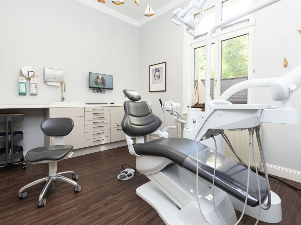 Moderner, freundlich heller Behandlungsraum einer Zahnarztpraxis mit ergonomischem Behandlungsstuhl, Assistenzhocker und moderner Behandlungseinheit vor einem großen Fenster sowie Schränken und Bildschirm im Hintergrund.
