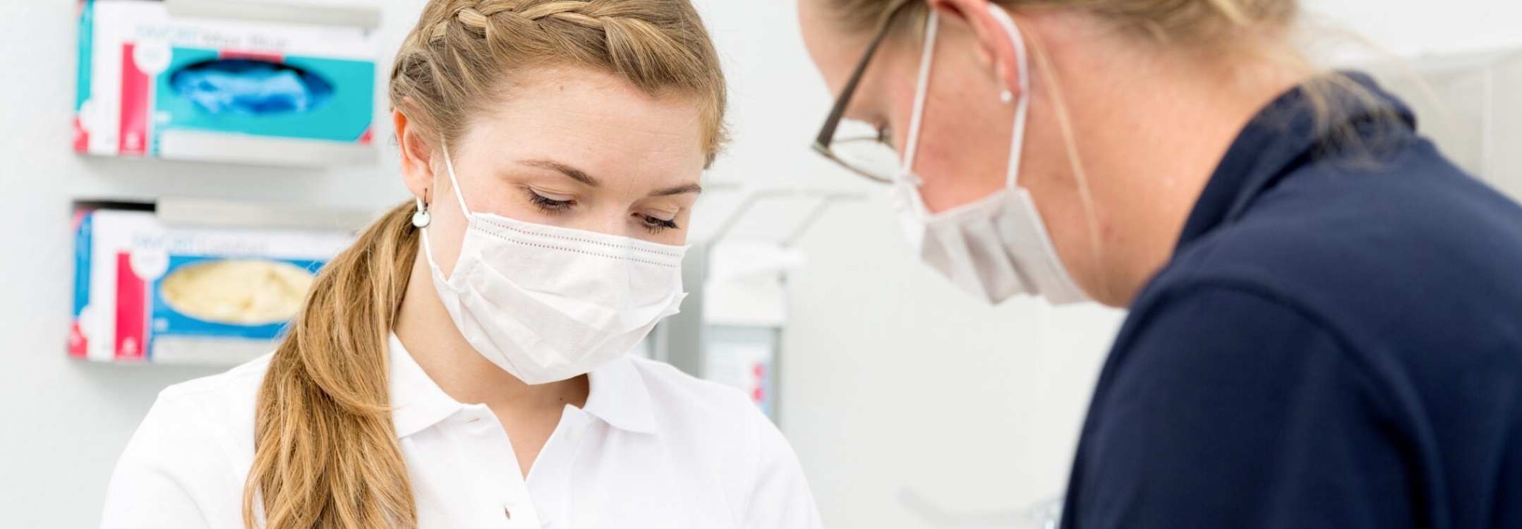 Intro Photo: Zwei zahnmedizinische Fachkräfte stehen nebeneinander in einer hellen Behandlungspraxis, tragen OP-Masken und Arbeitskleidung und beugen sich konzentriert über eine gerade durchzuführende Behandlung oder Vorbereitung eines Eingriffs.