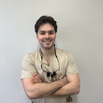 Porträt von Zahnarzt Michael Kapral aus DIE ZAHNARZTPRAXIS Stralsund, im beigen Kasack mit Lupenbrille um den Hals, vor hellem Hintergrund stehend und mit verschränkten Armen freundlich lächelnd.