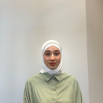 Porträt von Auszubildender Hawa Lorsanova aus DIE ZAHNARZTPRAXIS Stralsund, im grünen Kasack und weißem Kopftuch vor hellem Hintergrund, freundlich zur Kamera gewandt.